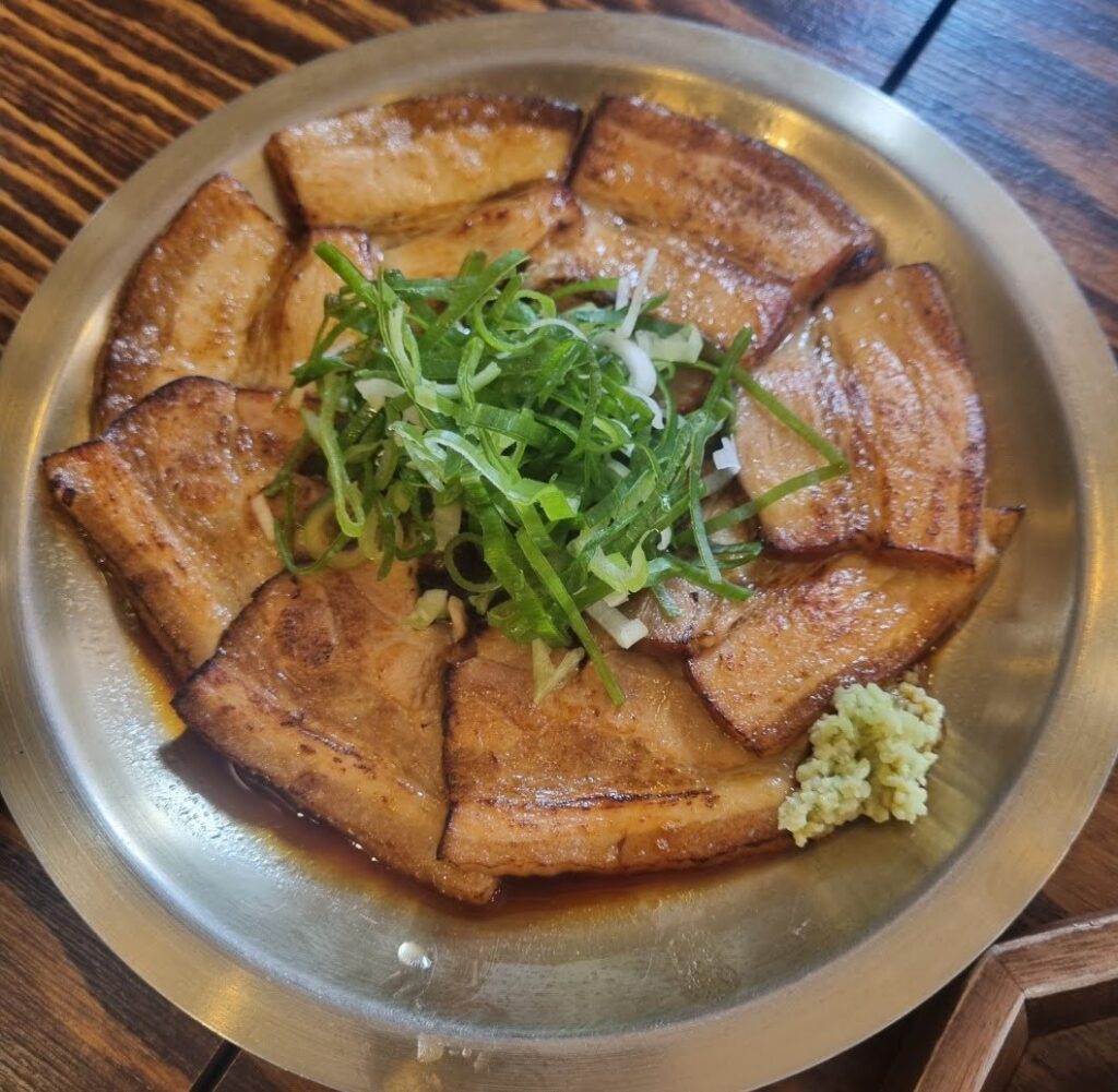 A plate of Oryukdon-Suyuk, Korean boiled pork belly slices arranged in a circular pattern, topped with green onions, and served with a side of wasabi.